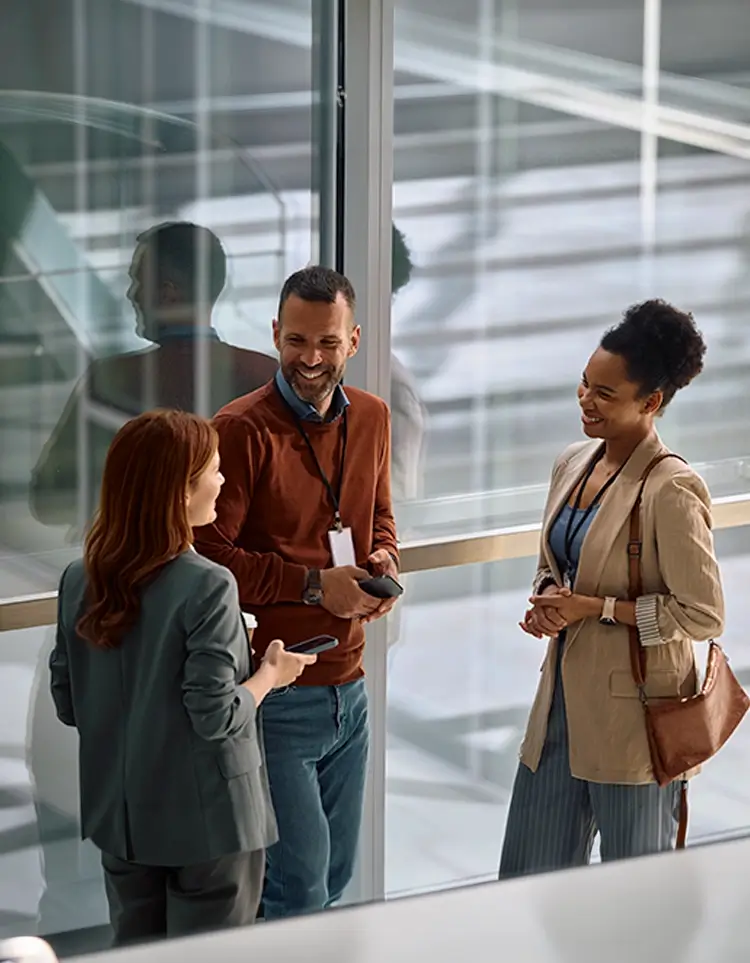 Three colleagues smiling and talking in a bright office hallway beside large windows