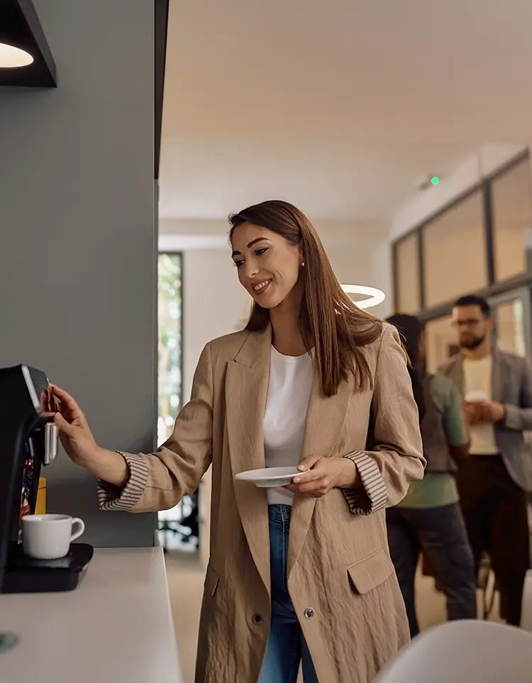 Woman using a coffee machine in an office kitchen while coworkers stand in the background