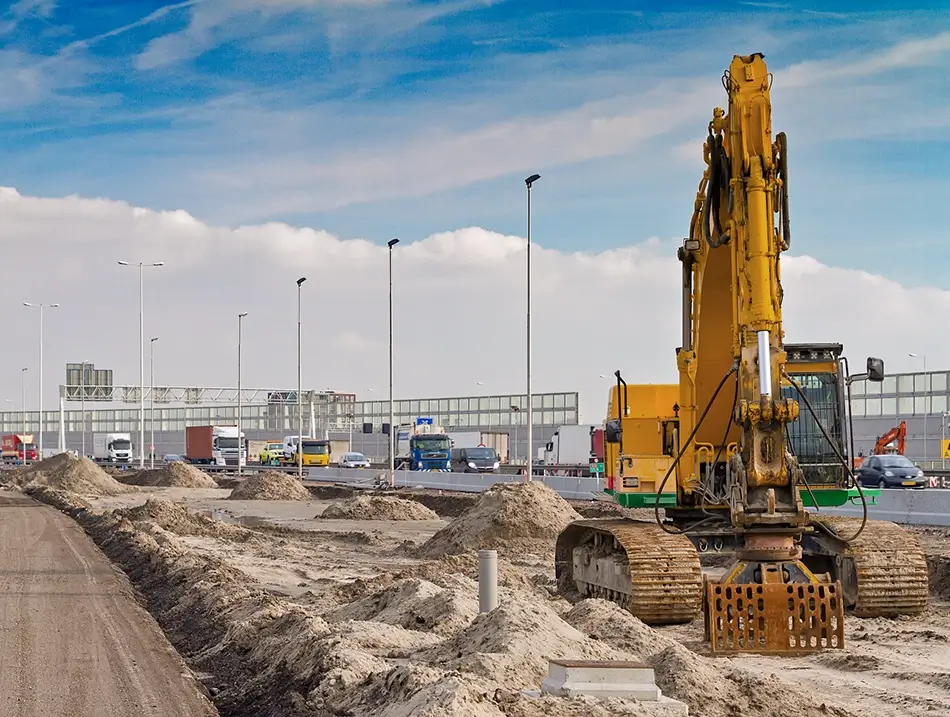 Yellow excavator at a road construction site with sand piles, machinery, and industrial buildings in the background