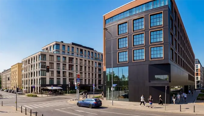 Clear daylight view of a modern urban street with contemporary buildings, pedestrians, a blue car, and a city intersection.