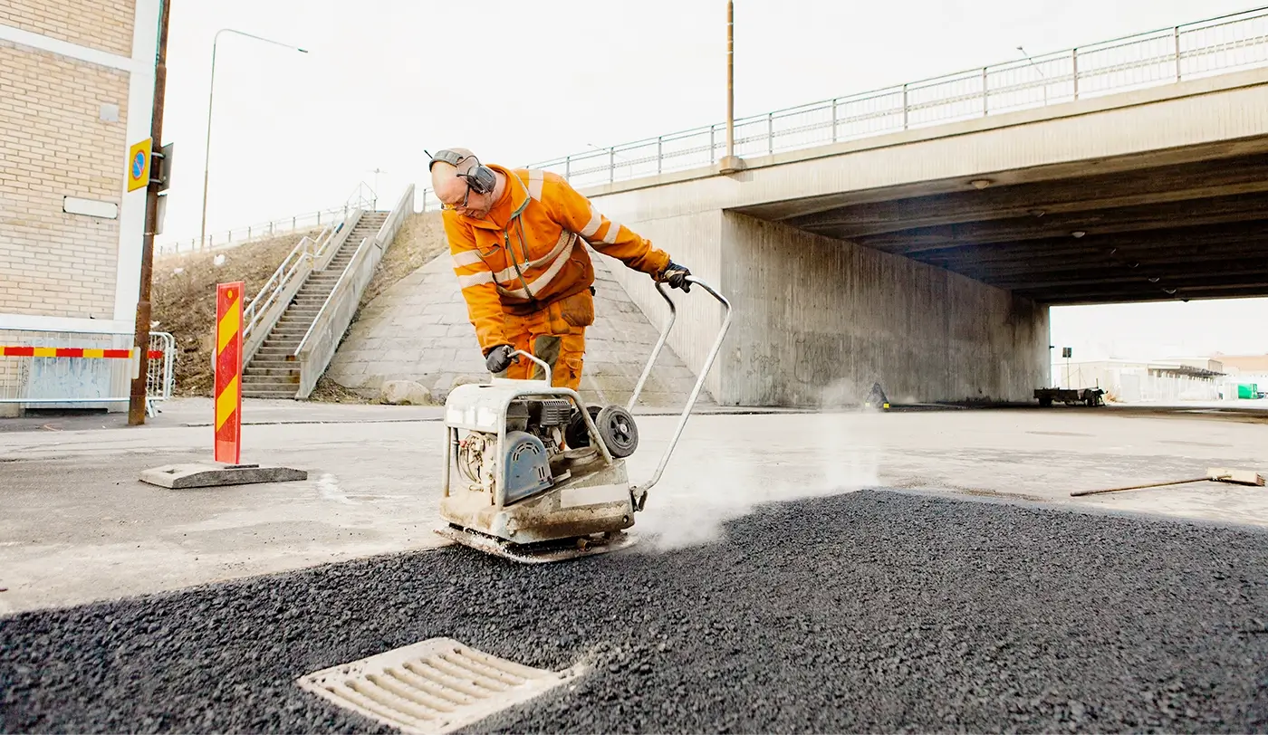 Road worker in high-visibility clothing compacting fresh asphalt near an underpass