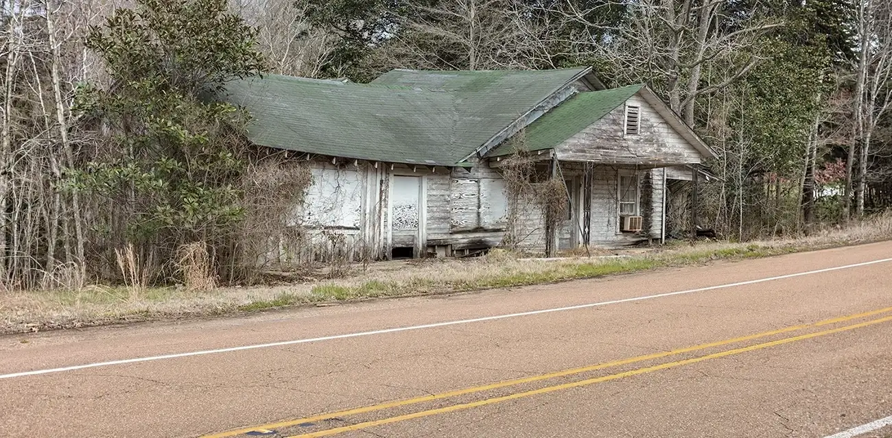 Weathered abandoned house beside a rural road with overgrown vegetation and a worn exterior