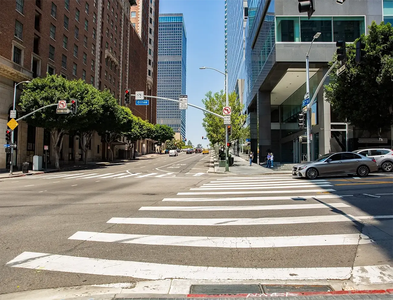 Downtown Los Angeles street intersection with crosswalk, traffic lights, office buildings, and parked cars