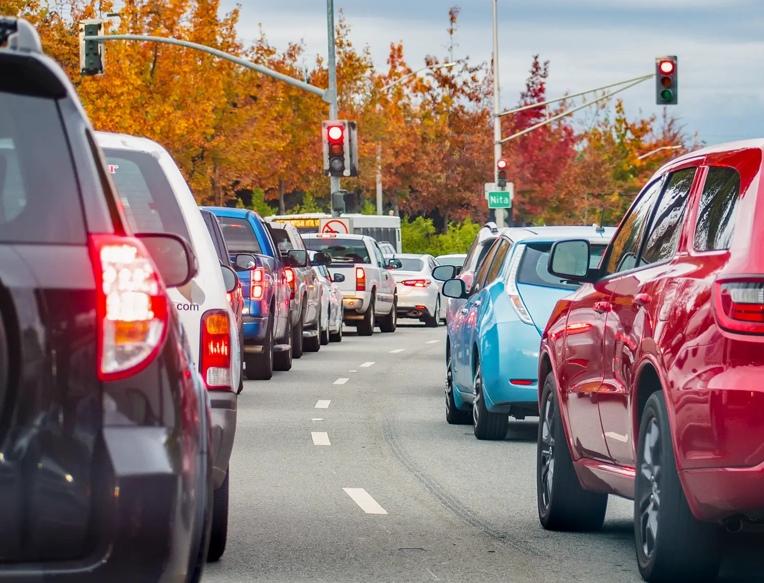 Cars lined up at a red traffic light on a busy urban road with autumn trees in the background