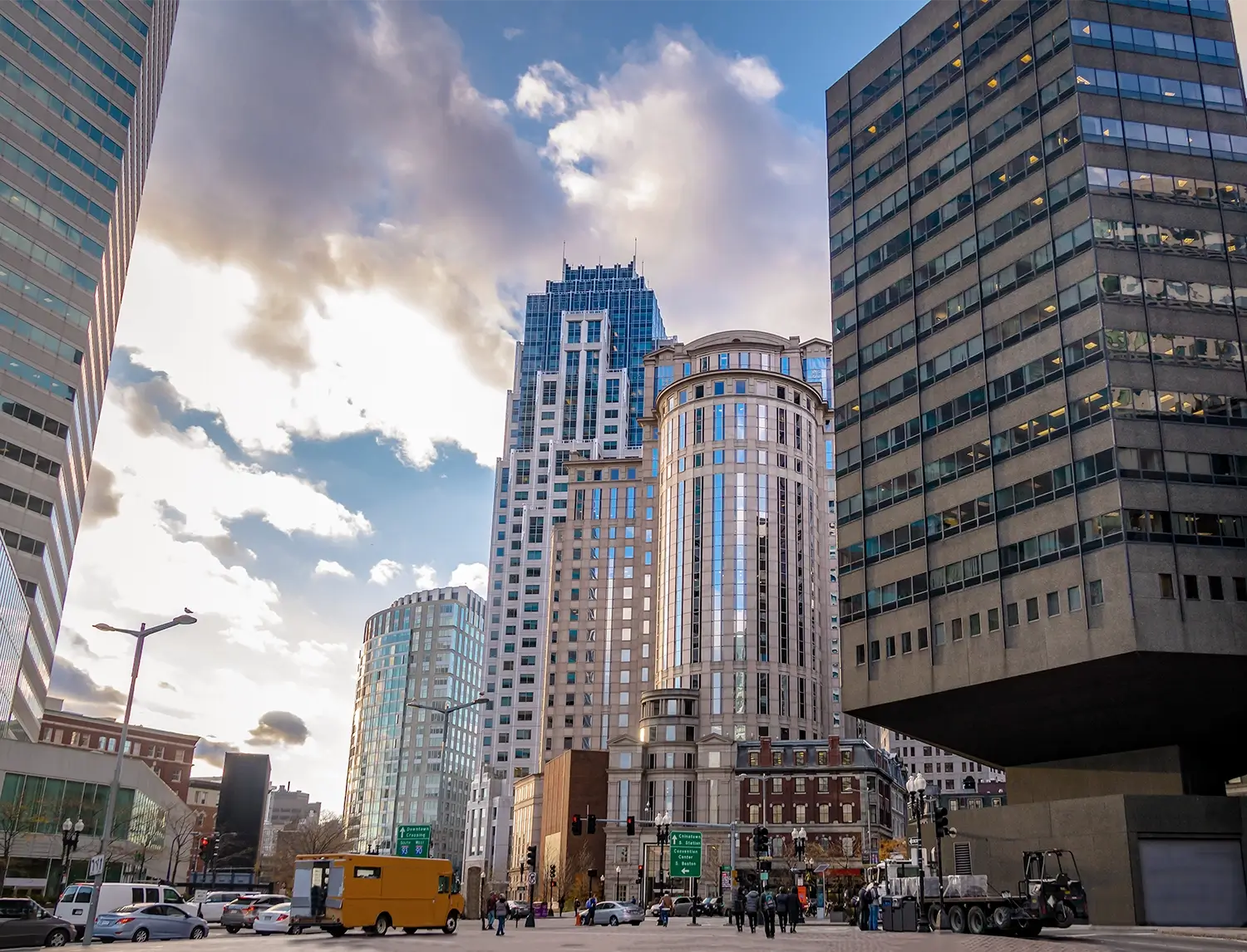 Downtown Boston street scene with tall office buildings, traffic, pedestrians, and a cloudy sky