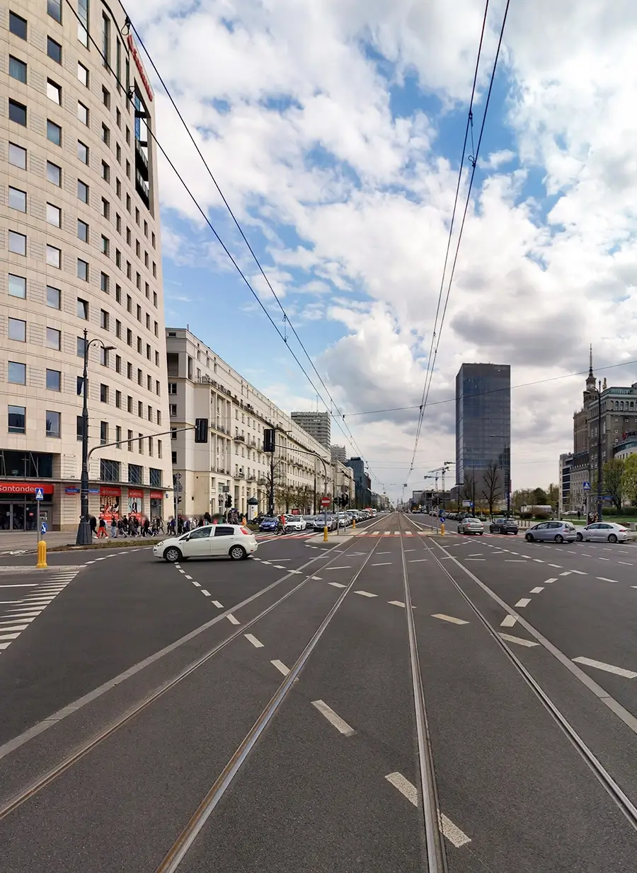 Wide urban boulevard with tram tracks, lane markings, surrounding buildings, and traffic under a partly cloudy sky