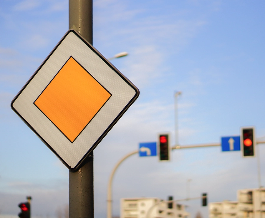 Close-up of a priority road sign on a pole with traffic lights and road direction signs in the background
