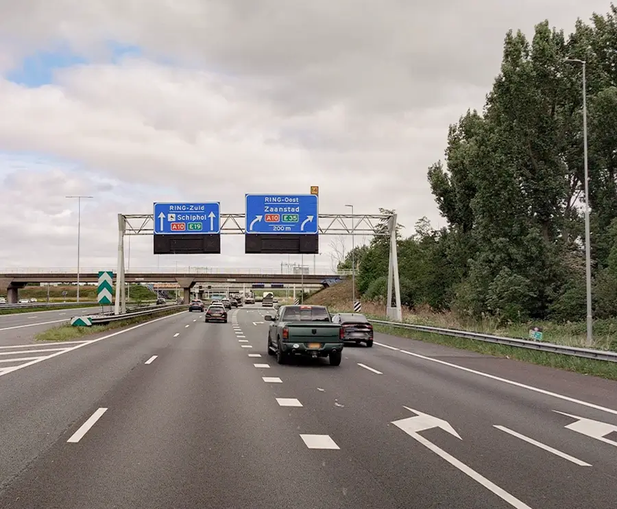 Multi-lane highway with directional overhead signs for Ring-Zuid, Ring-Oost, Schiphol, and Zaanstad under a cloudy sky