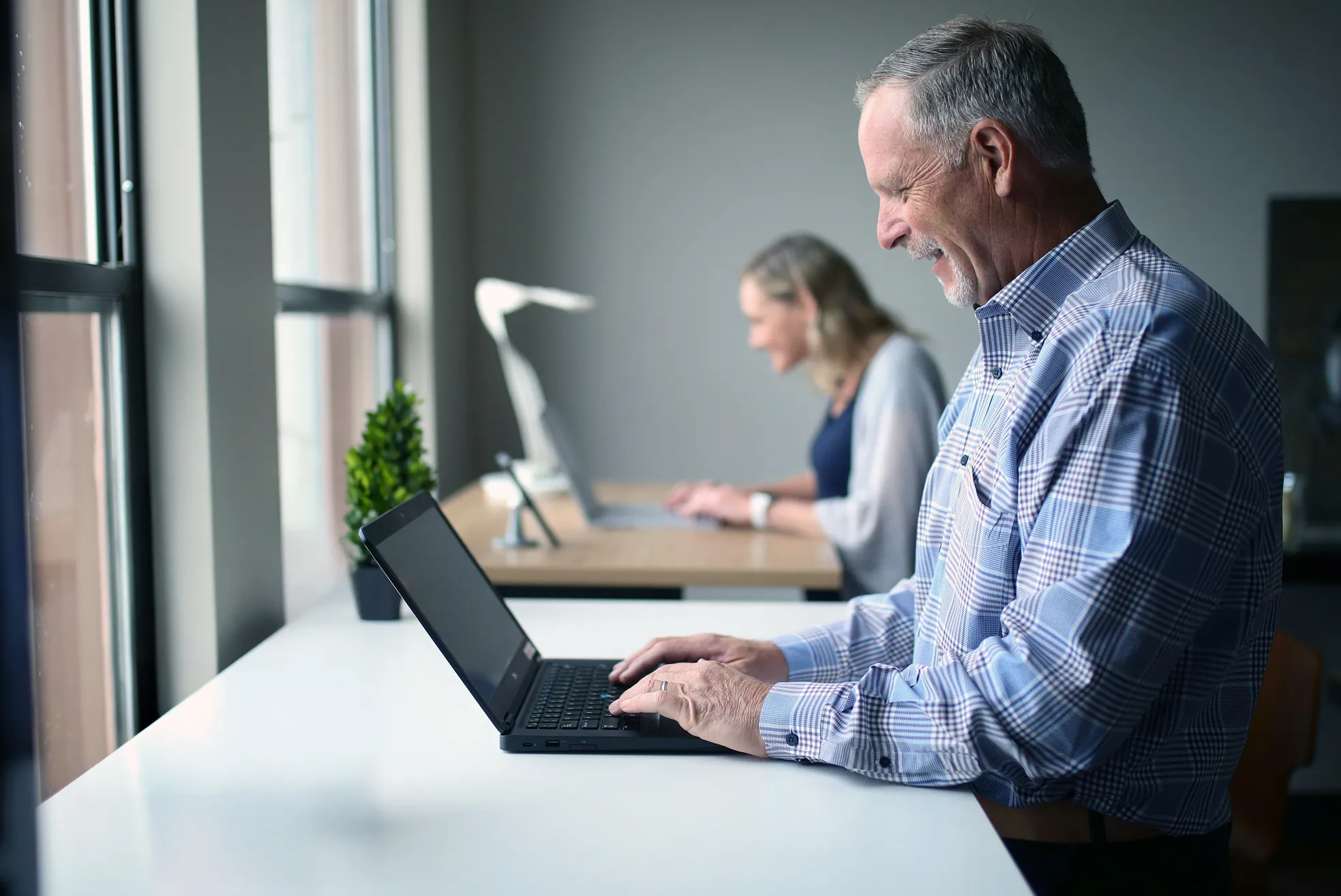 Smiling older man and woman working on laptops while sitting at desks by a window in a modern office.