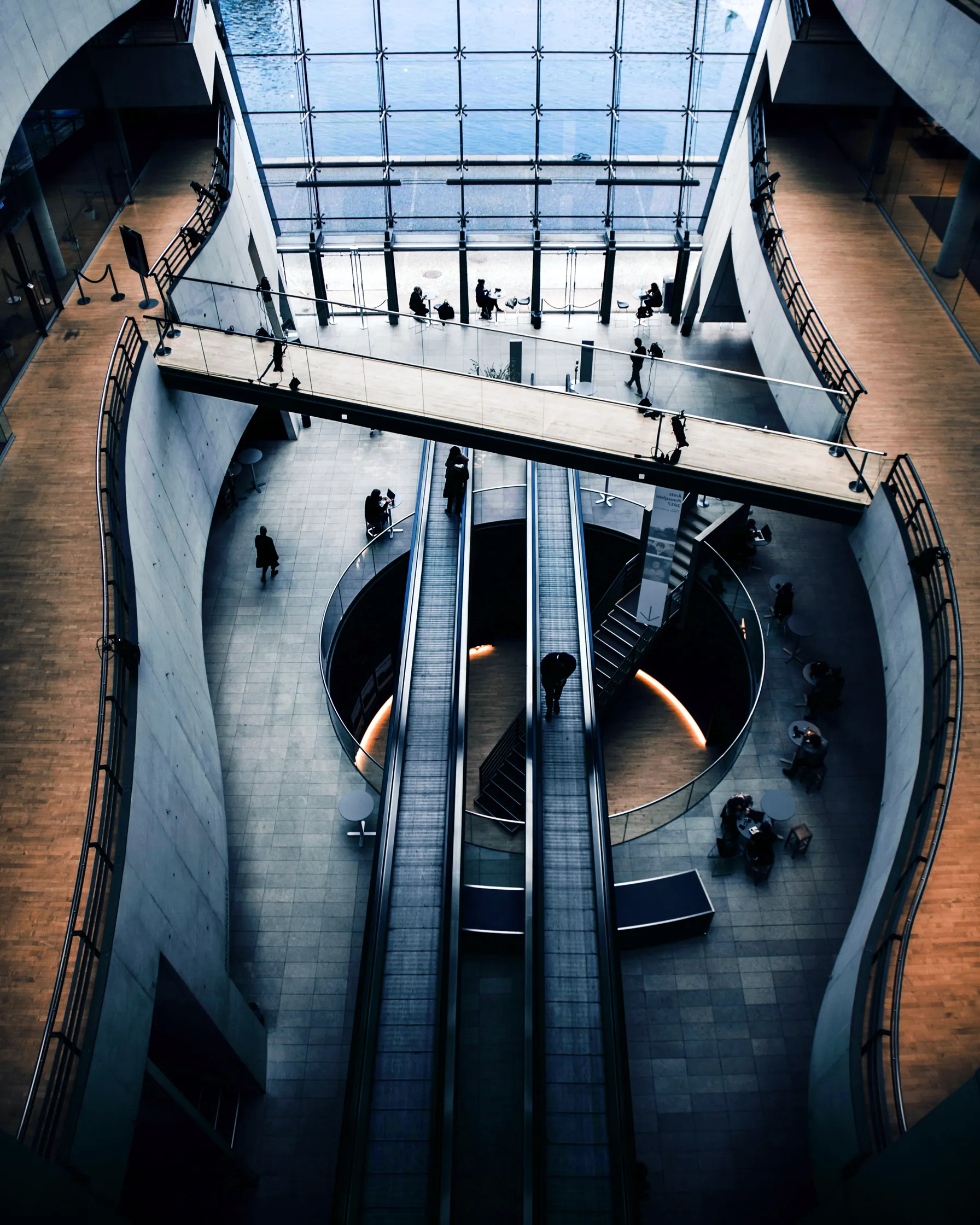 Interior view of a modern building with two moving walkways, a curved wooden bridge above, and people walking or sitting near large glass windows.