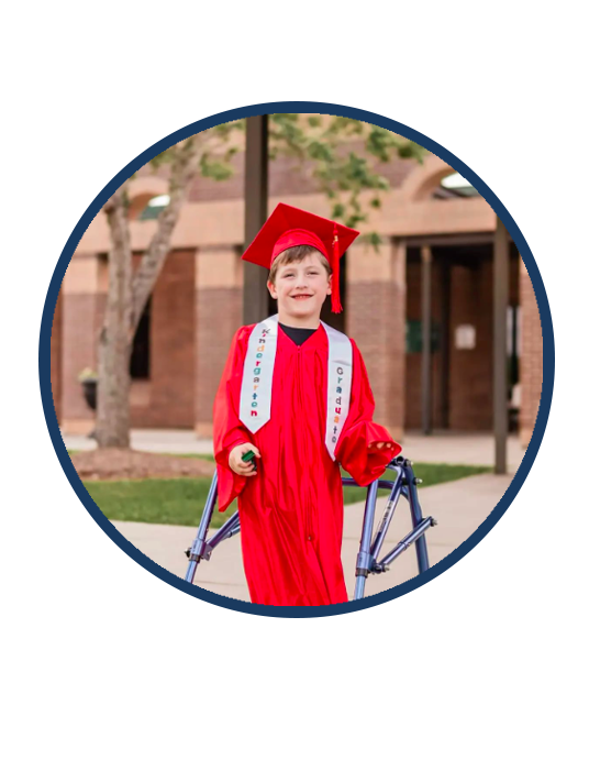 Young boy in a red graduation gown and cap using a walker outside a building.