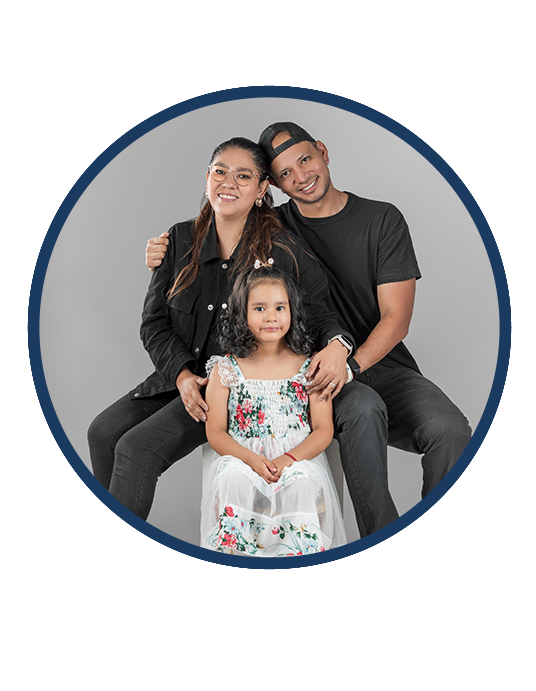 Smiling mother and father in black with daughter in white floral dress sitting in front of them.
