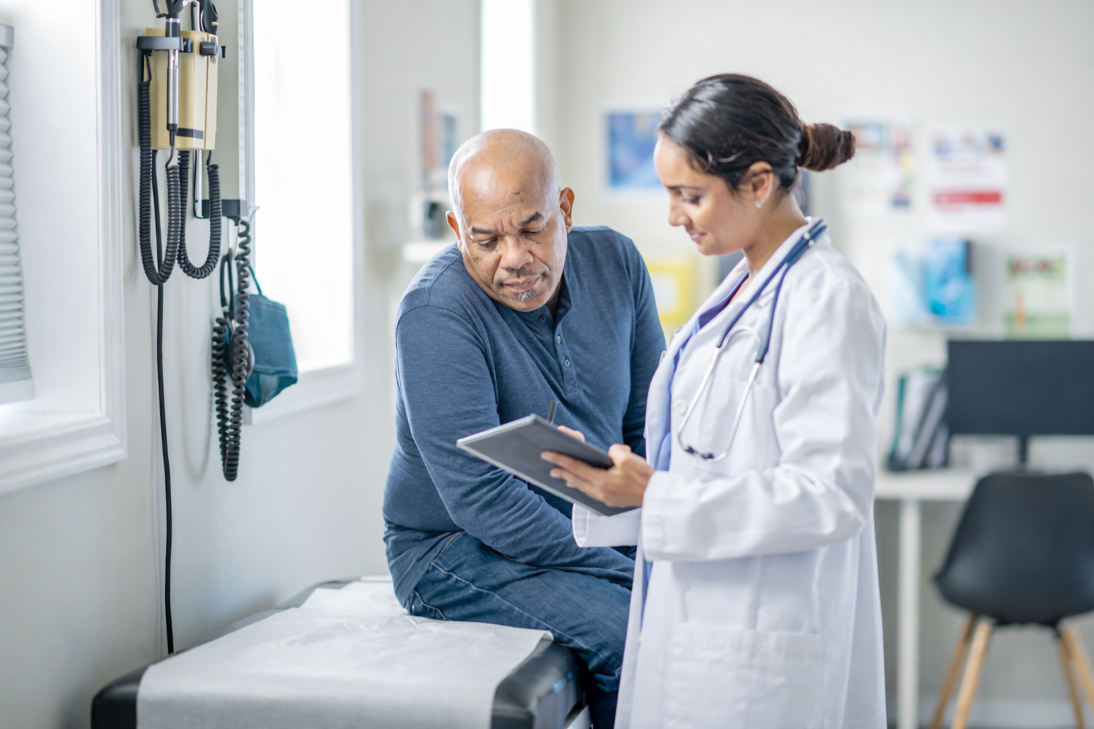 Doctor in white coat writing on clipboard while patient sits on exam table in medical office.