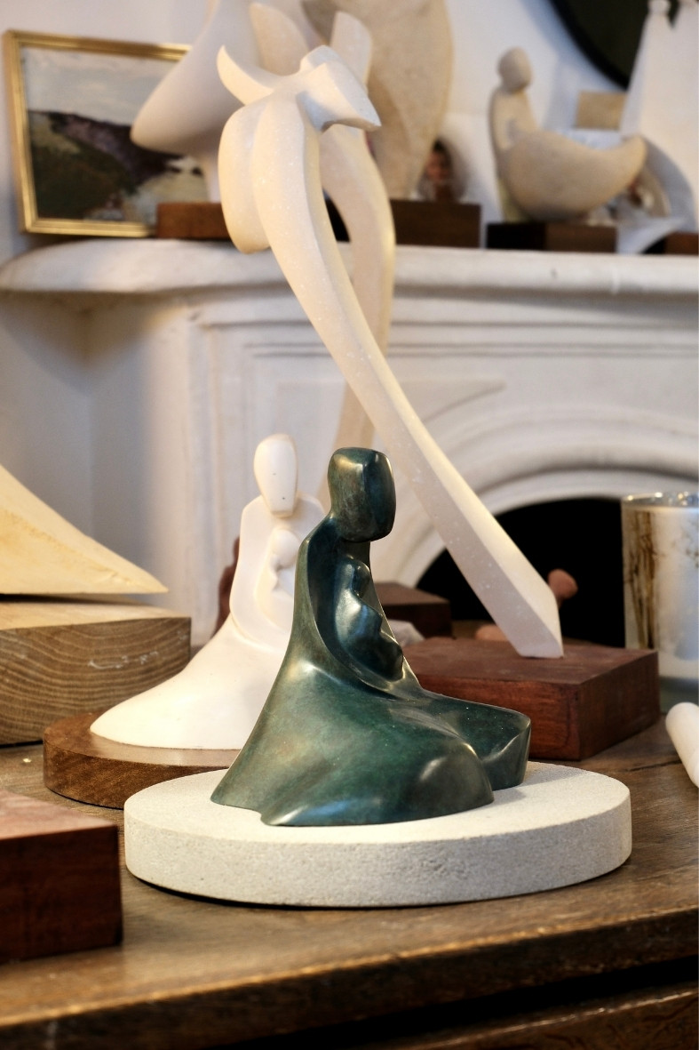 Sculpture Sculpture Vierge de Tendresse de l'Atelier Philippe Gancel en bronze représentant une figure humaine assise tenant un bébé dans un atelier avec d'autres sculptures et matériaux en arrière-plan.