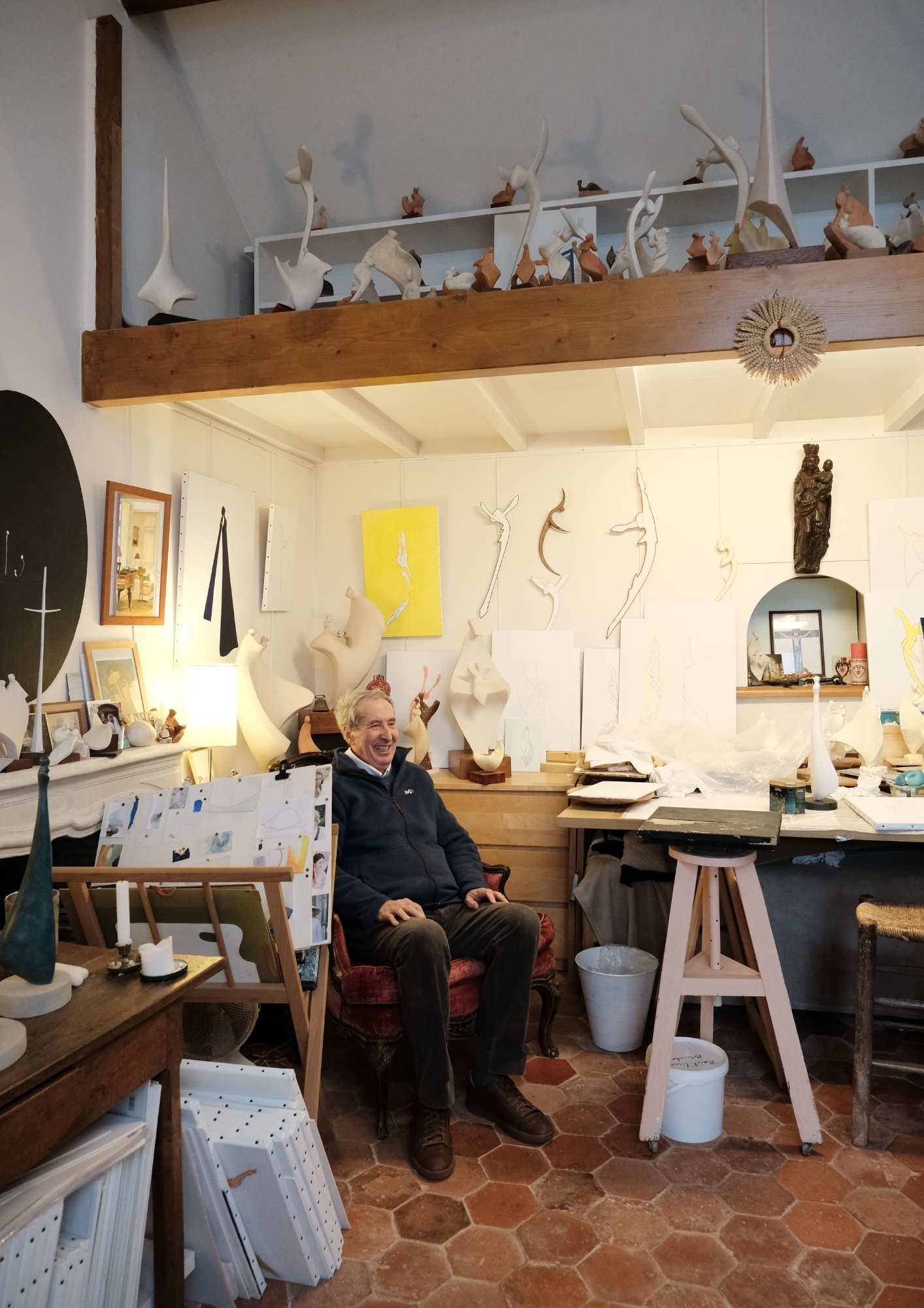 L'artiste sculpteur Philippe Gancel assis dans un fauteuil rouge dans son atelier lumineux, entouré de sculptures modernes et de dessins accrochés aux murs.