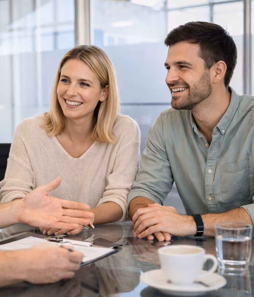 Ein lächelndes junges Paar sitzt an einem Glastisch und unterhält sich mit einer Person, die ein Klemmbrett in der Hand hält.