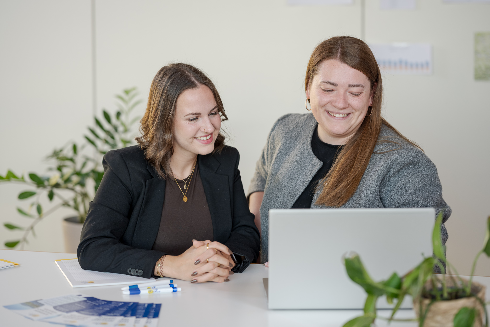Two women sitting at a table, smiling and looking at a laptop screen in a bright office.