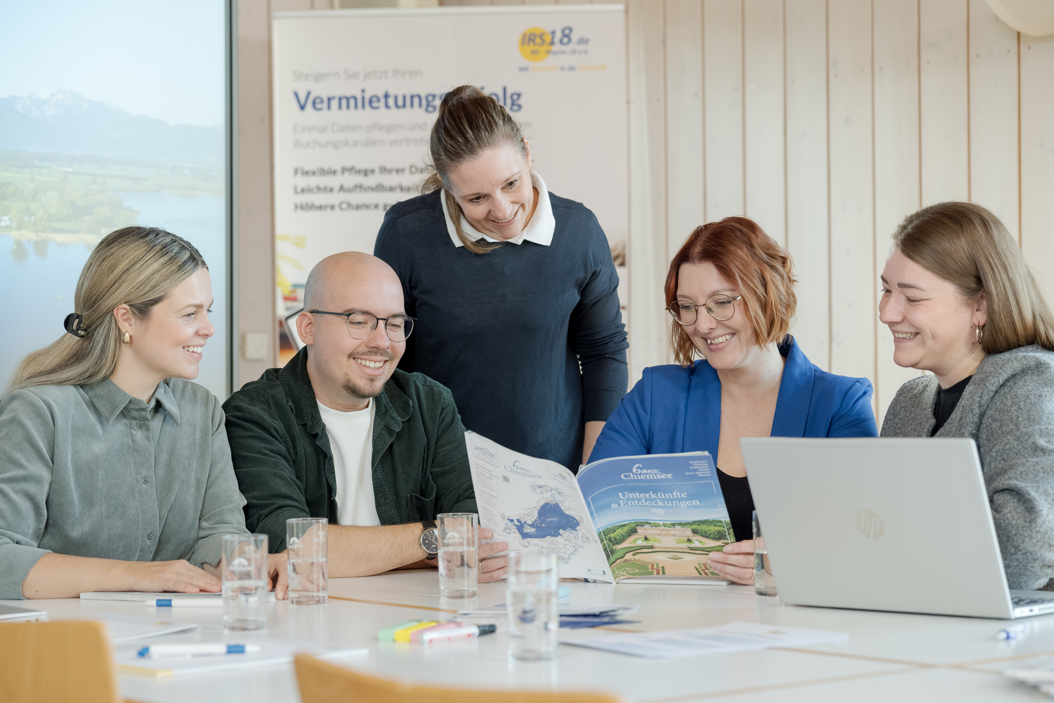 Five colleagues gathered around a table smiling and looking at a brochure together, with a laptop and glasses of water on the table.