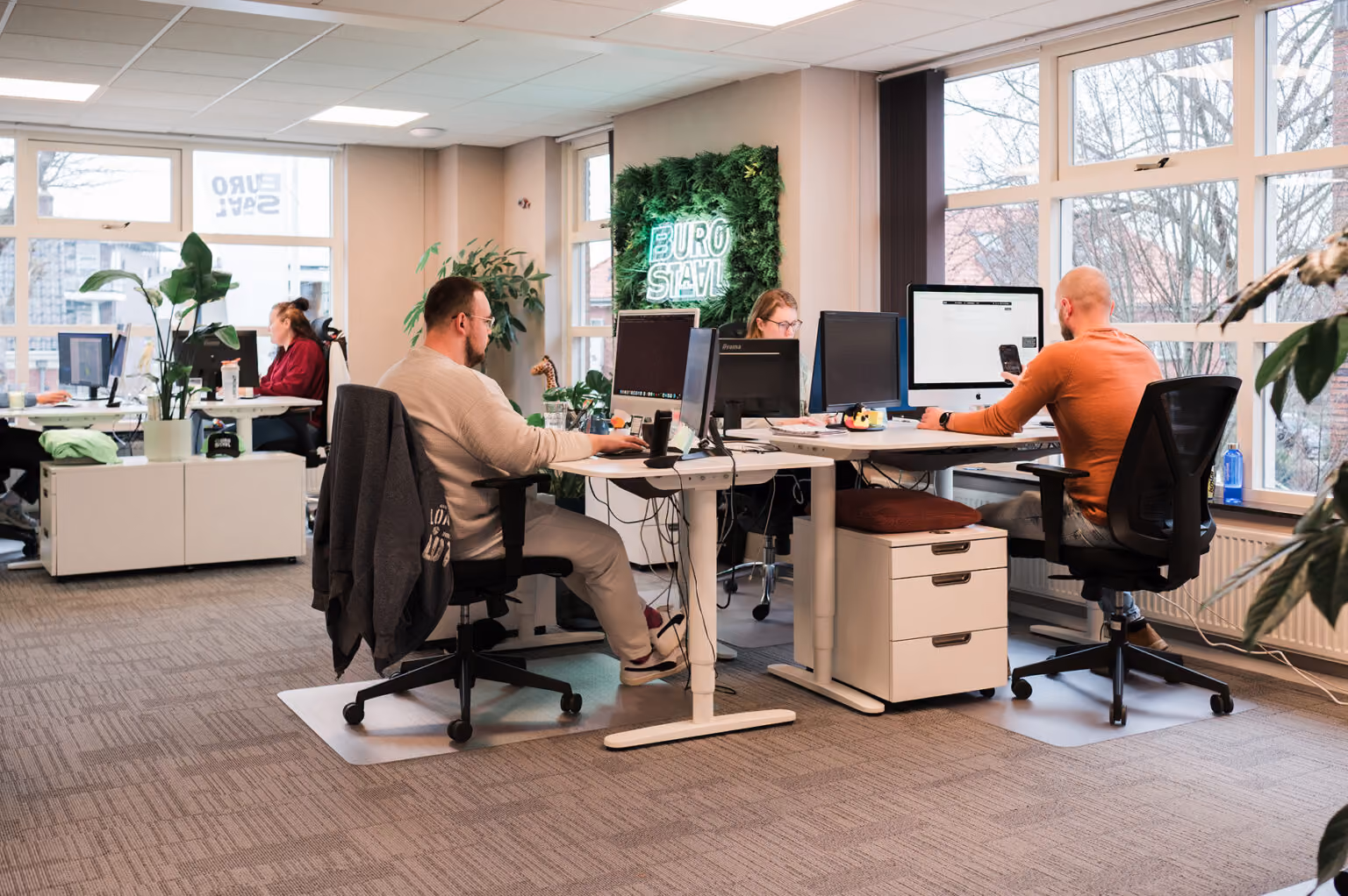 Mensen werken aan computers in een modern kantoor met grote ramen en planten.