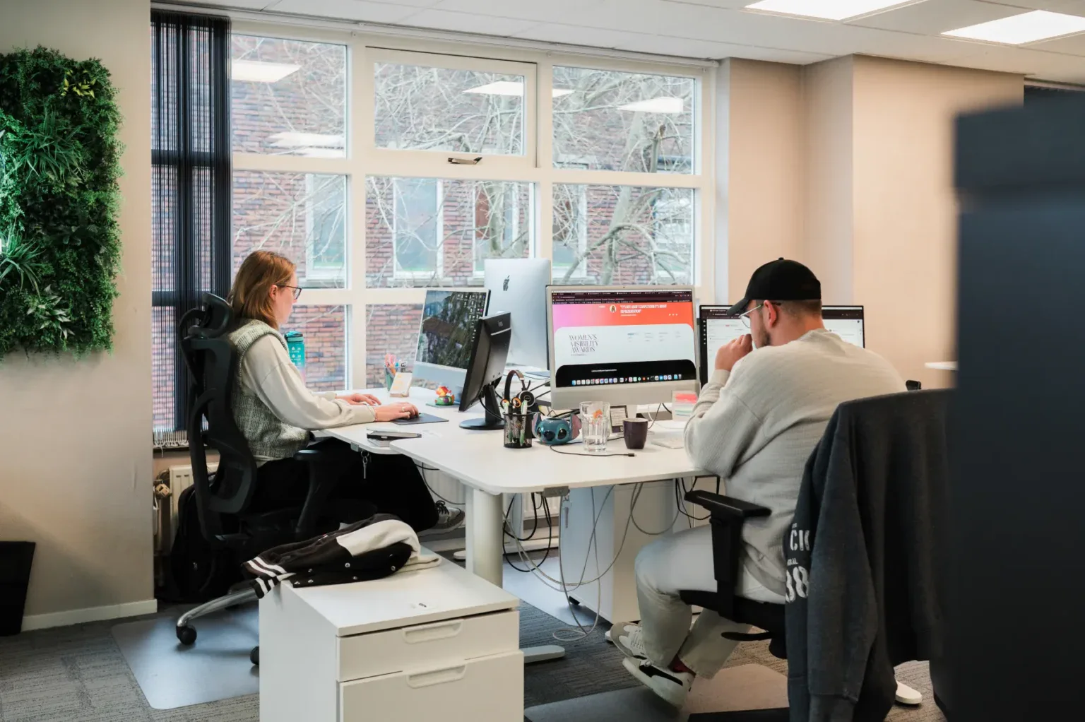 Twee mensen werken op computers aan een gedeeld bureau in een modern kantoor met grote ramen.