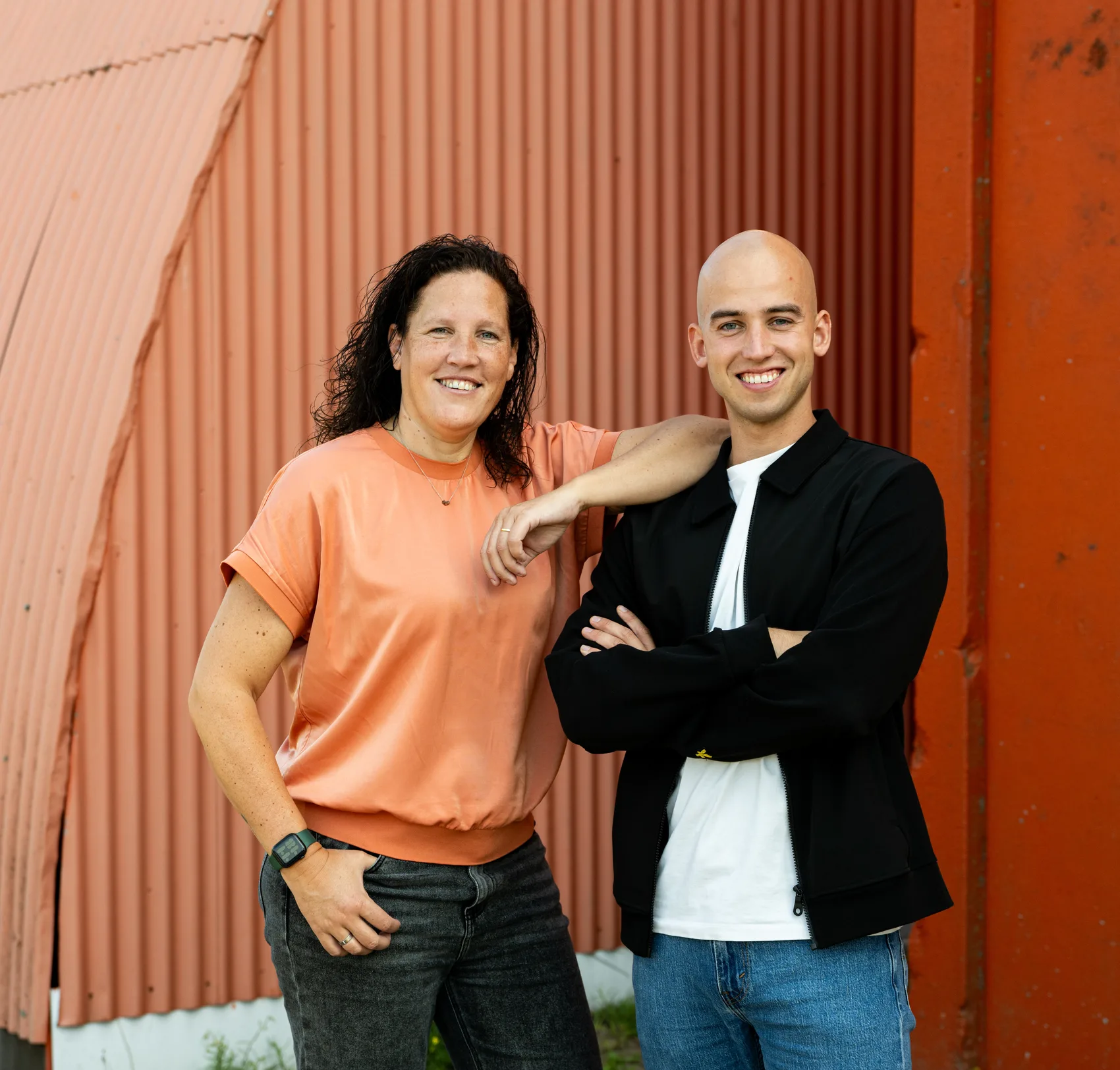Twee collega's van Buro Staal staan lachend naast elkaar voor een rode golfplaatwand
