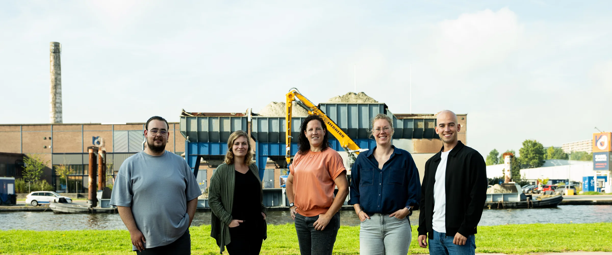 Het Buro Staal team van vijf staat buiten aan het water, met een industrieterrein en gele kraan op de achtergrond