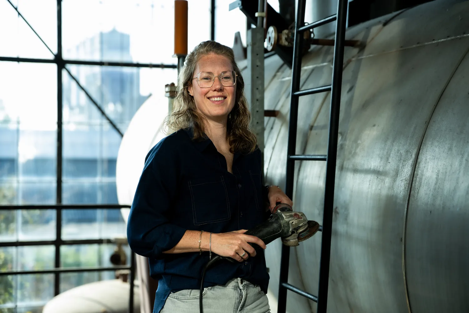 Rosanne in een donkerblauw overhemd met een haakse slijper bij een stalen ketel.