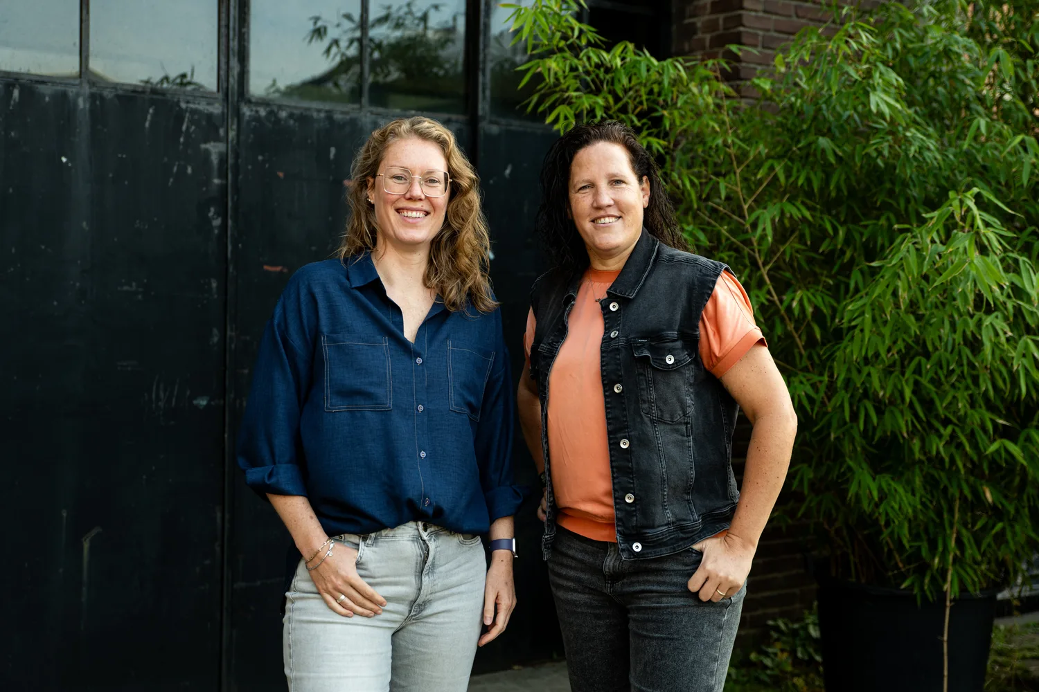 Annemieke en Rosanne staan lachend naast elkaar voor een donkere deur met bamboe.