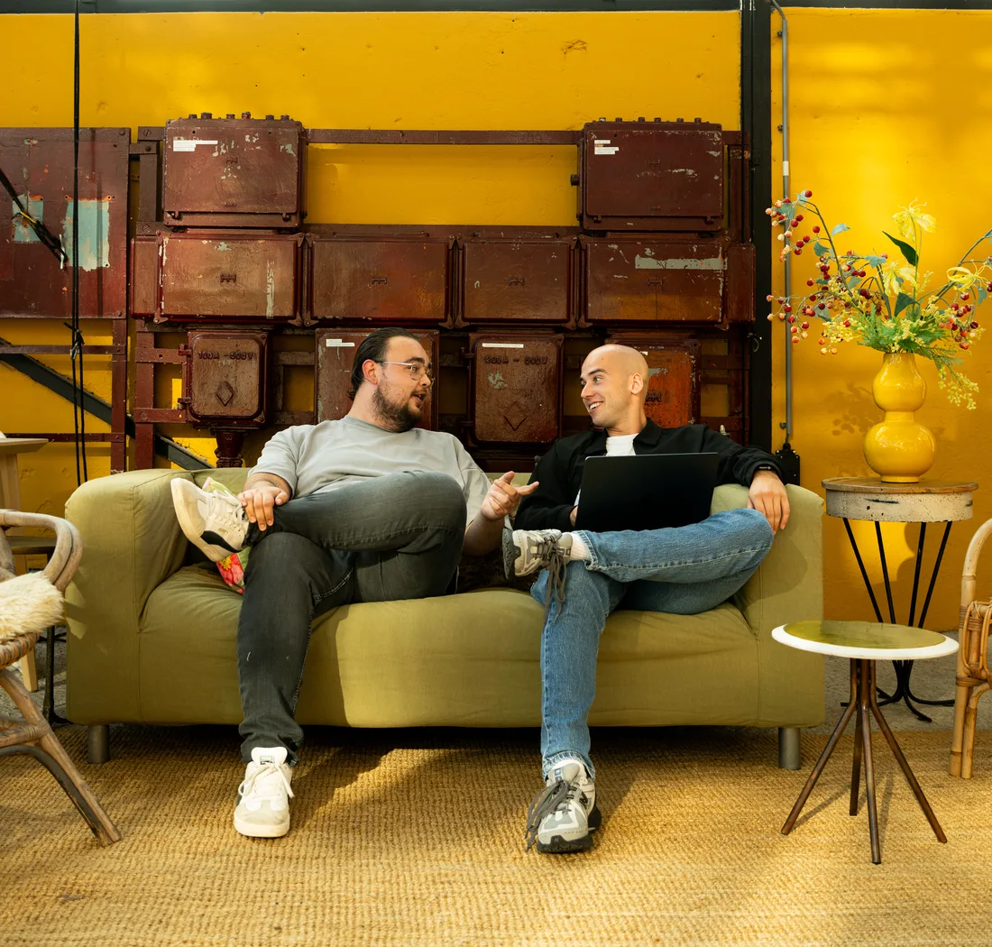 Coen en Kevin zitten op de groene bank in de gele lounge en praten met elkaar.