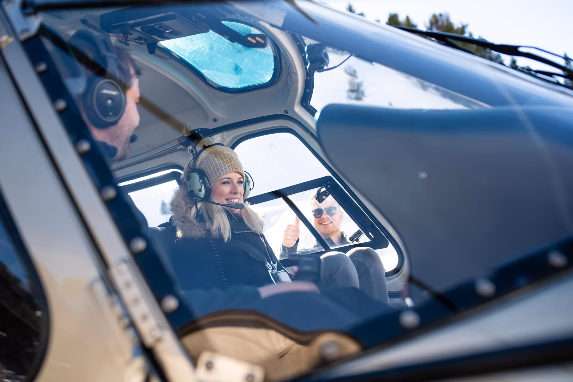 Two people inside a helicopter cockpit, one woman wearing a winter hat and headset smiling, and a man outside giving a thumbs up.
