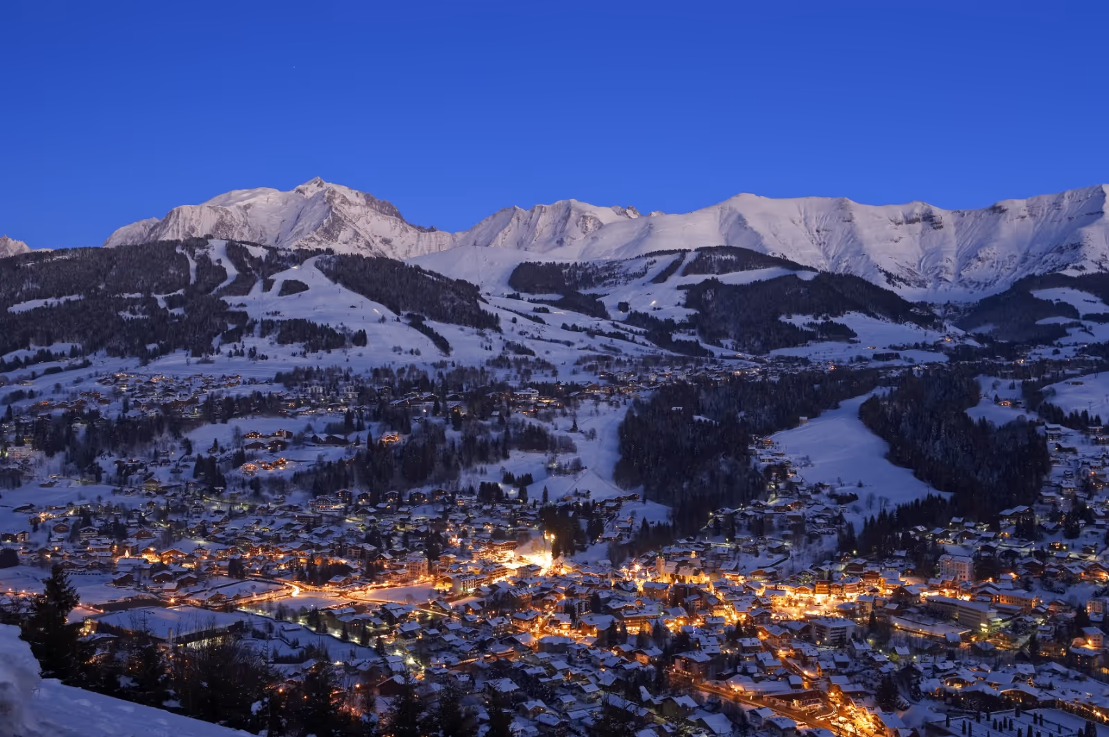 Snow-covered mountain village at dusk with illuminated buildings and clear blue sky.