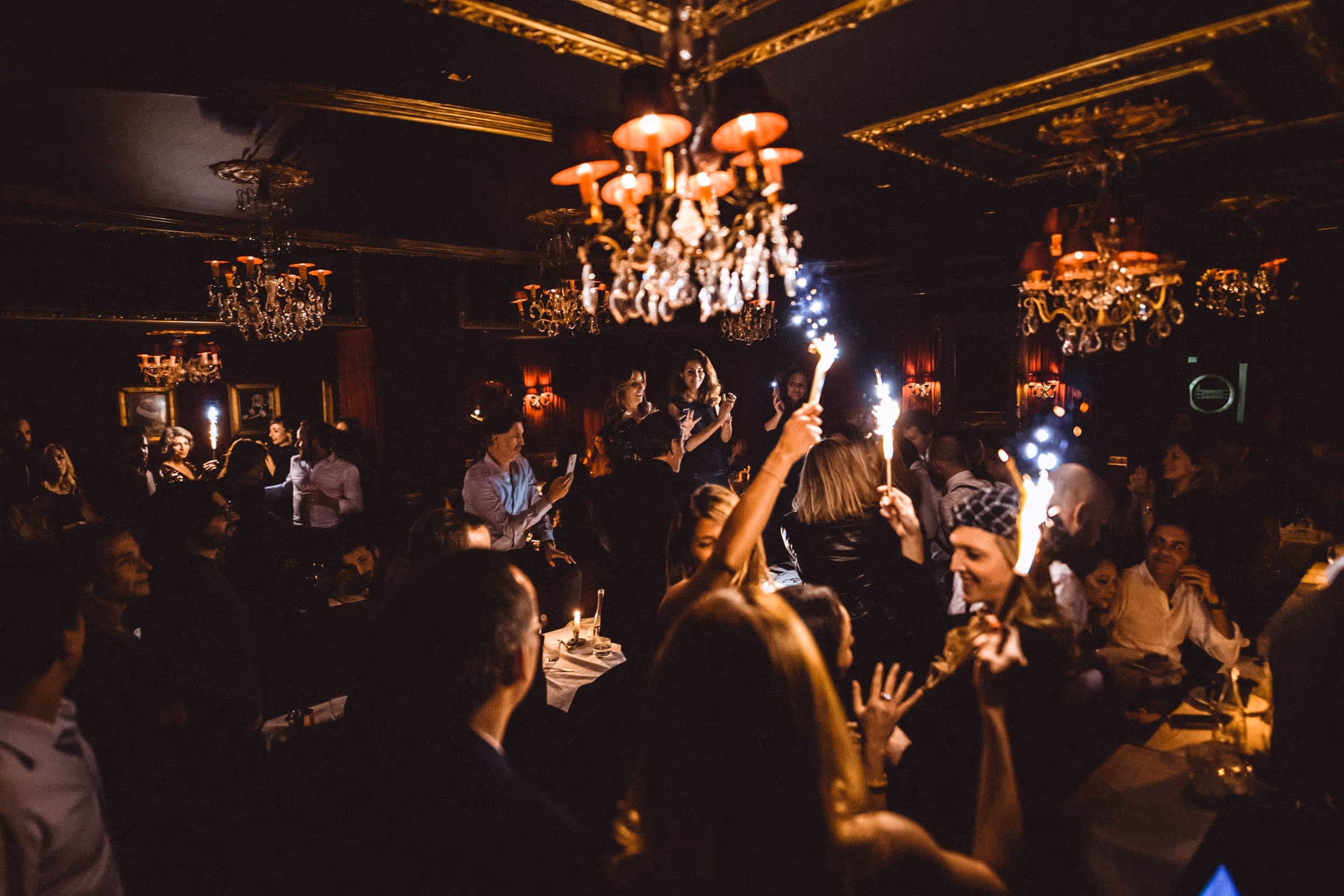 Crowded dimly lit room with people holding sparklers celebrating under ornate chandeliers.