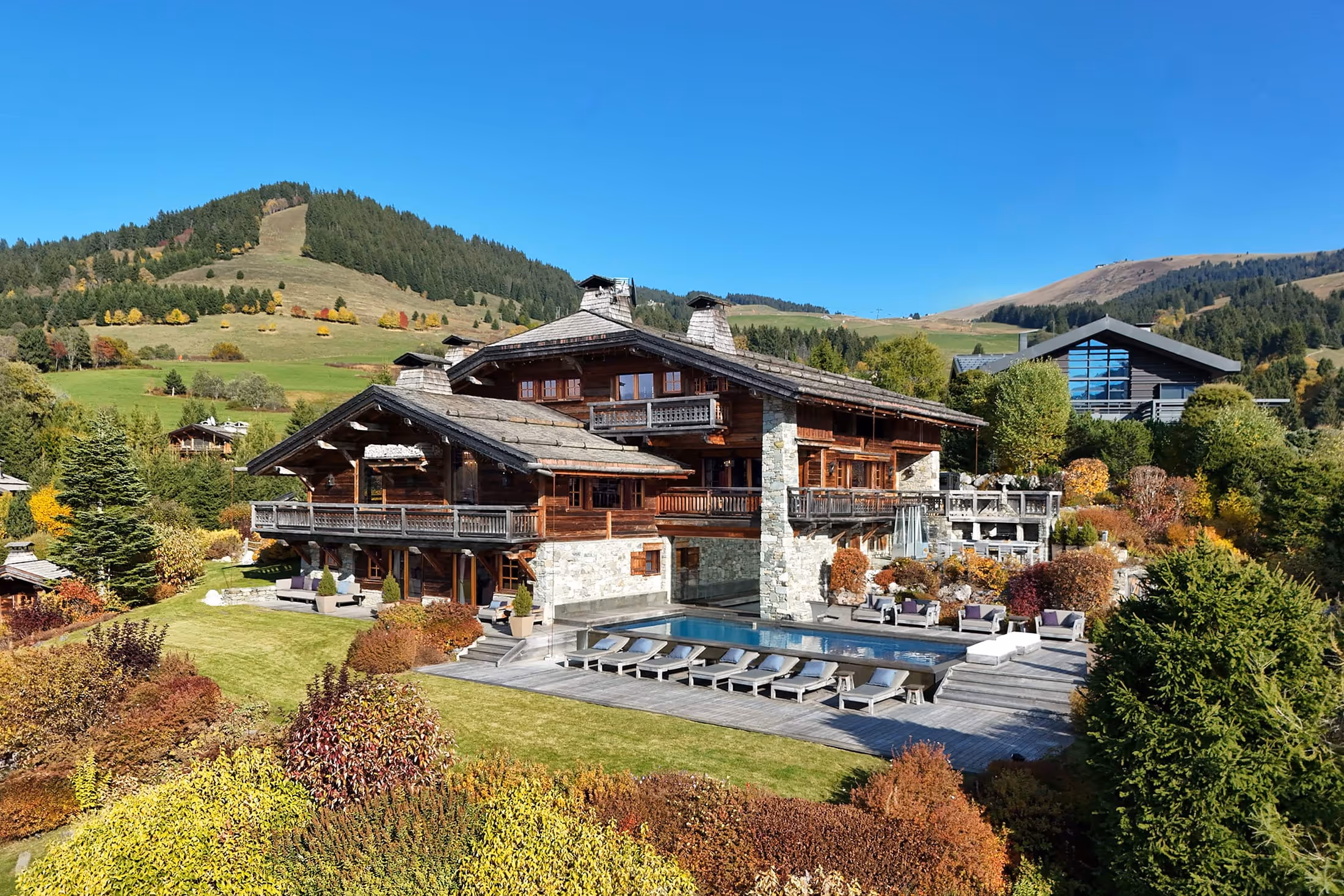 Large wooden chalet Berne in Megeve with stone accents surrounded by landscaped gardens, lounge chairs, and a swimming pool, set against green hills and clear blue sky.