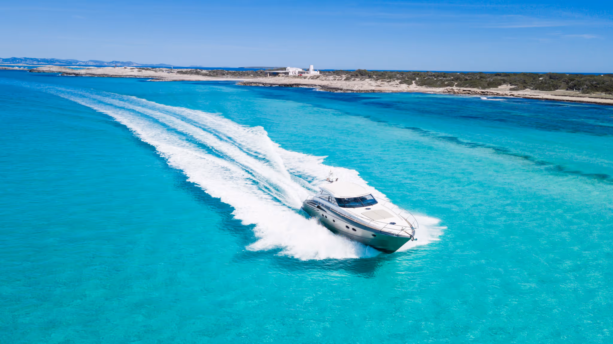 White motor yacht speeding through clear turquoise water near a sandy shoreline with sparse vegetation.