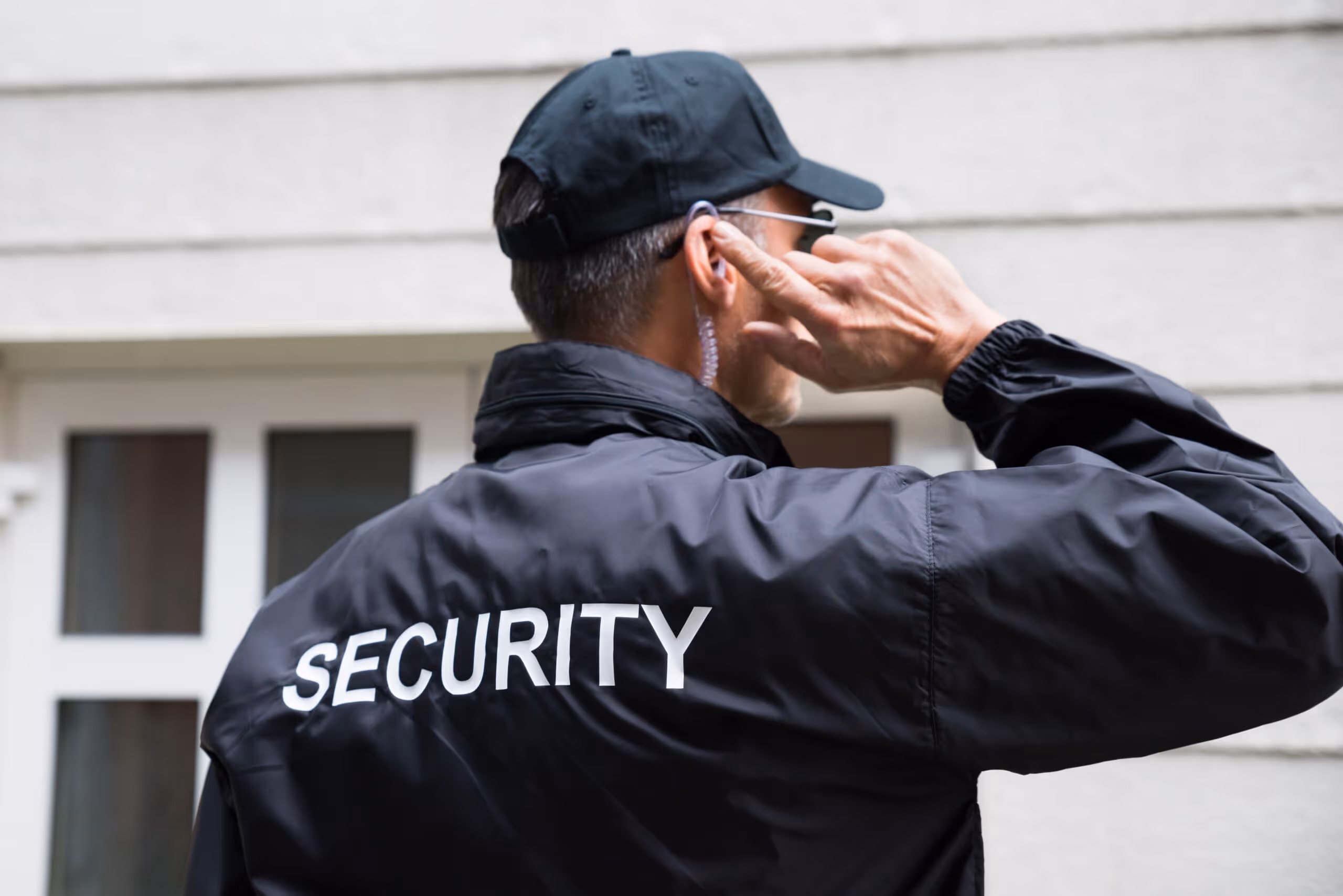 Security guard in black jacket and cap adjusting earpiece near a building entrance.
