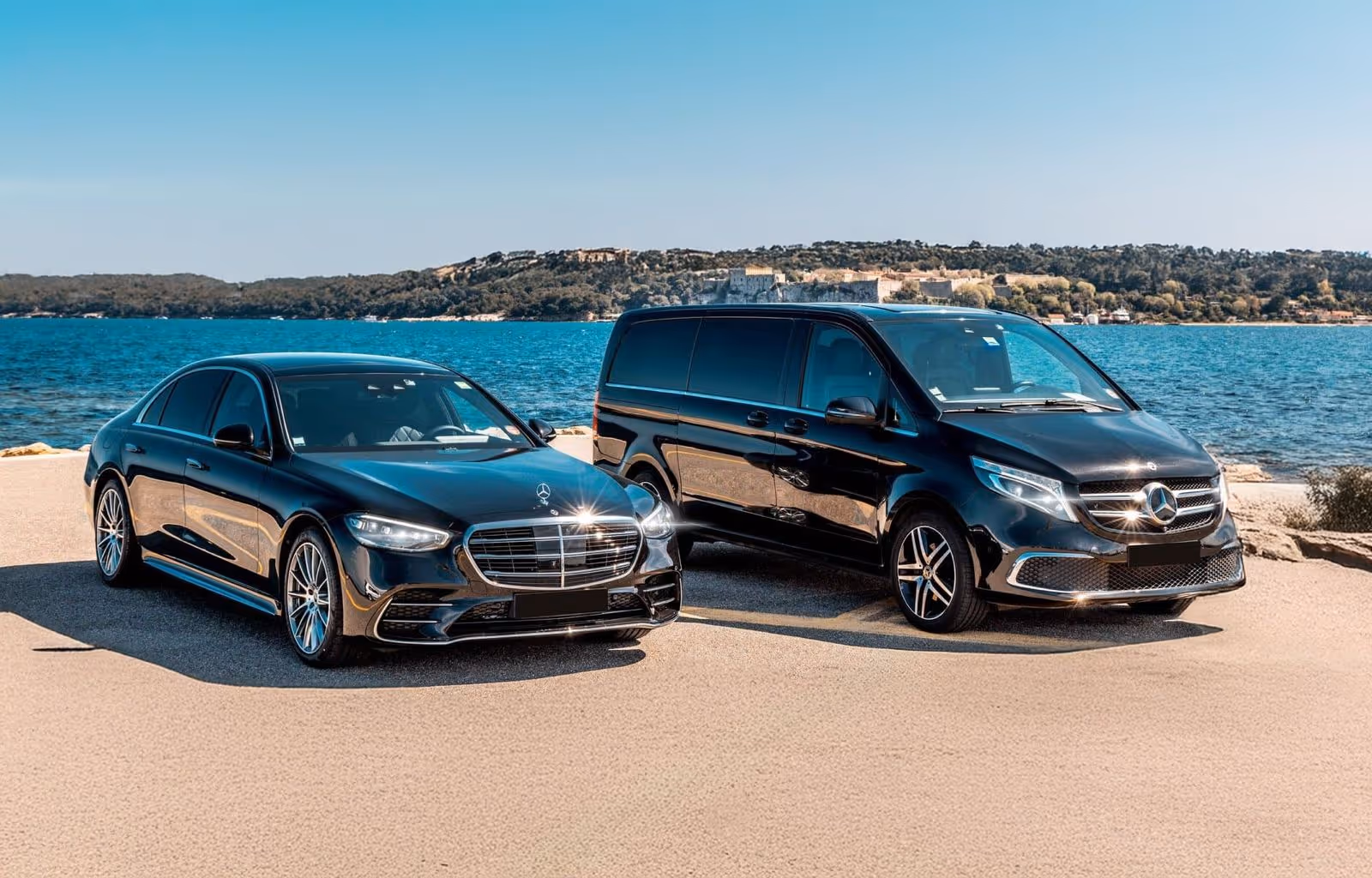 Two black Mercedes-Benz vehicles, a sedan and a van, parked near a waterfront with a clear blue sky.