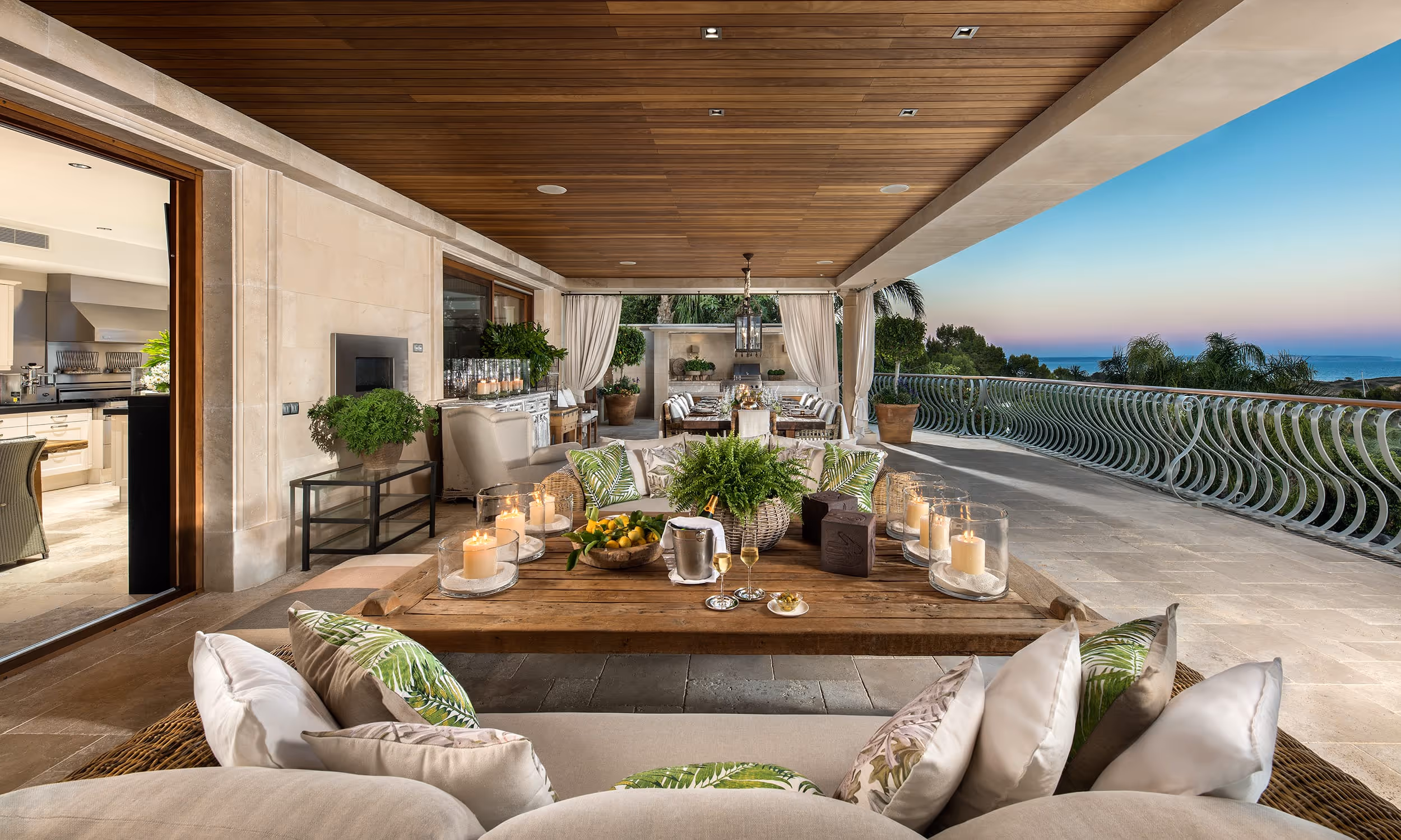 Spacious covered outdoor terrace with comfortable wicker seating, wooden coffee table with candles and plants, dining area, and ocean view at sunset.