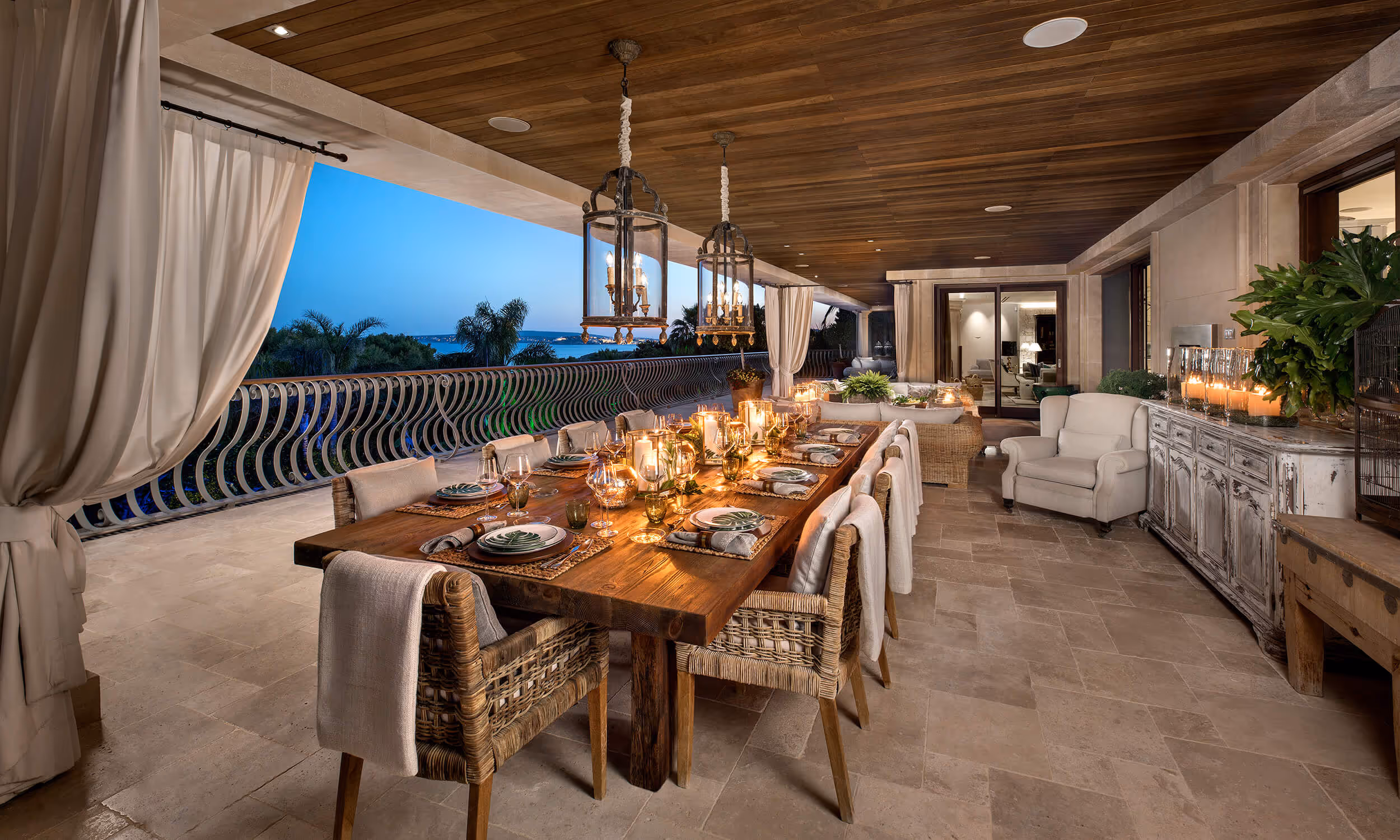 Elegant outdoor dining area with a wooden table set for twelve under hanging lanterns, with sea view at dusk.