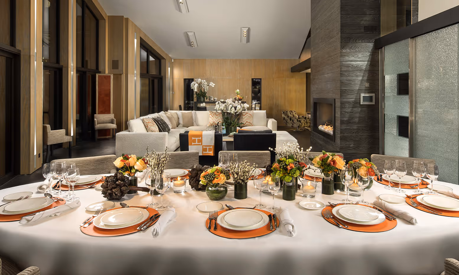 Elegant dining table set with white tablecloth, orange placemats, glassware, and floral centerpieces in a modern living room with beige sofa and a fireplace.