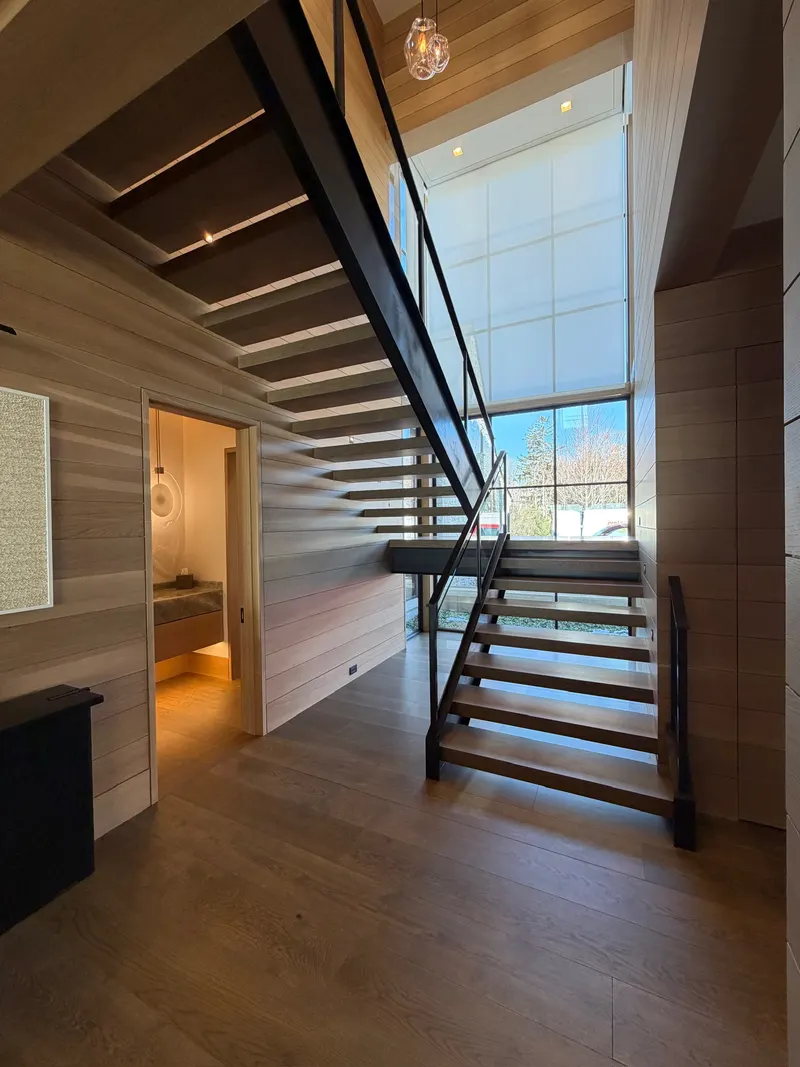 Modern interior with wooden flooring and walls, featuring a floating staircase with black railing and large window bringing natural light.