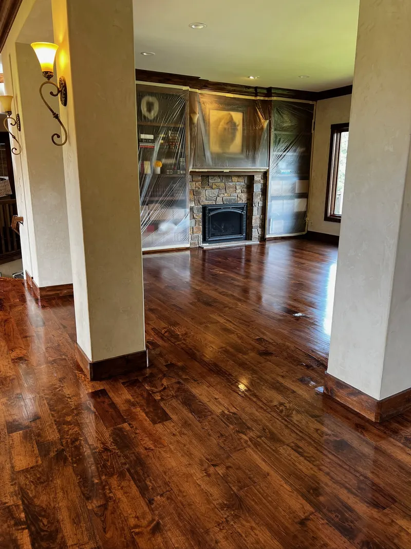 Empty living room with polished dark hardwood floors, two beige pillars, stone fireplace, and bookshelves covered in plastic.