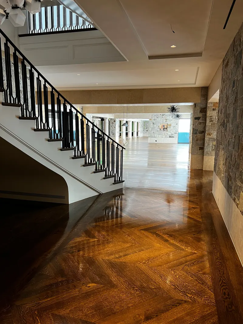 Interior view of a spacious room with polished wooden herringbone floor, stone walls, and a staircase with black railing.