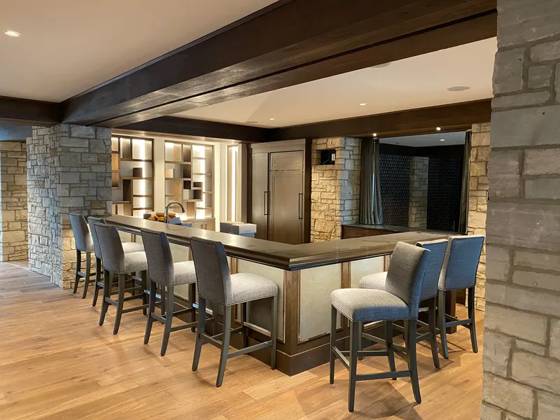 L-shaped home bar with gray cushioned bar stools, stone columns, wooden floor, and soft ambient lighting.