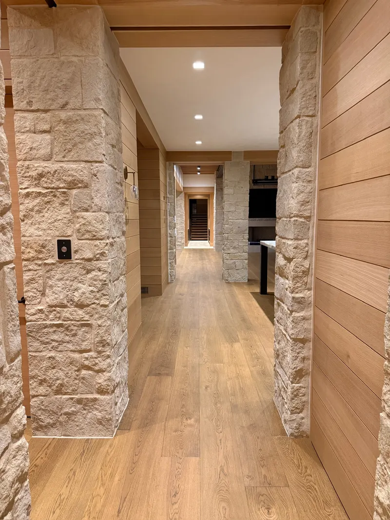 Hallway with wide plank oak flooring, rough stone columns, and horizontal wooden shiplap walls under recessed ceiling lights.