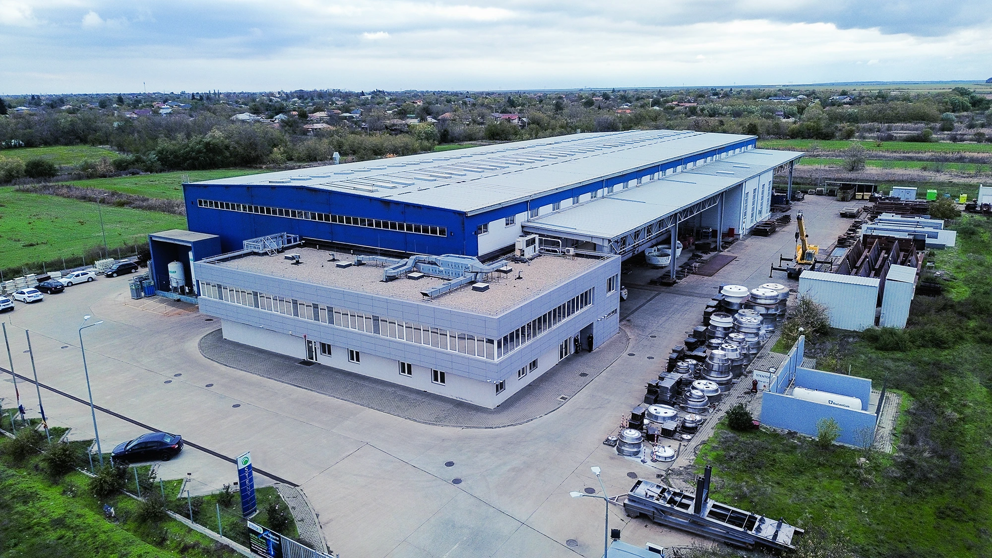 Aerial view of an industrial building with a blue upper section and surrounded by metal components and vehicles in a rural area.