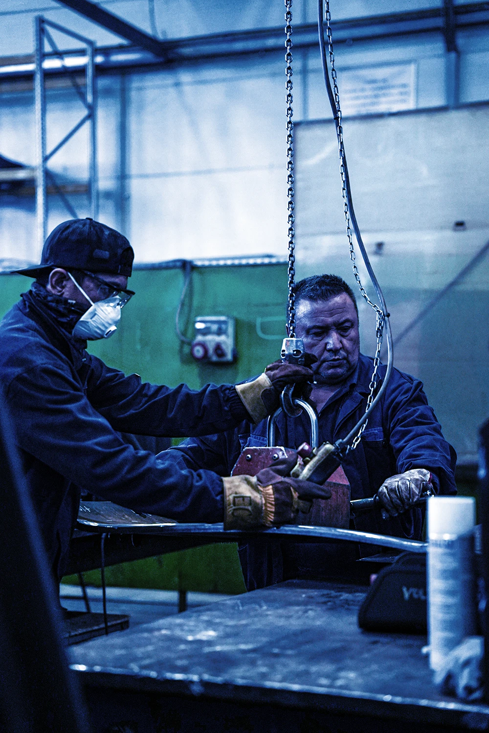 Doi muncitori în atelier metalurgic manipulează o bucată grea de metal cu ajutorul unui cârlig suspendat de o lanț.