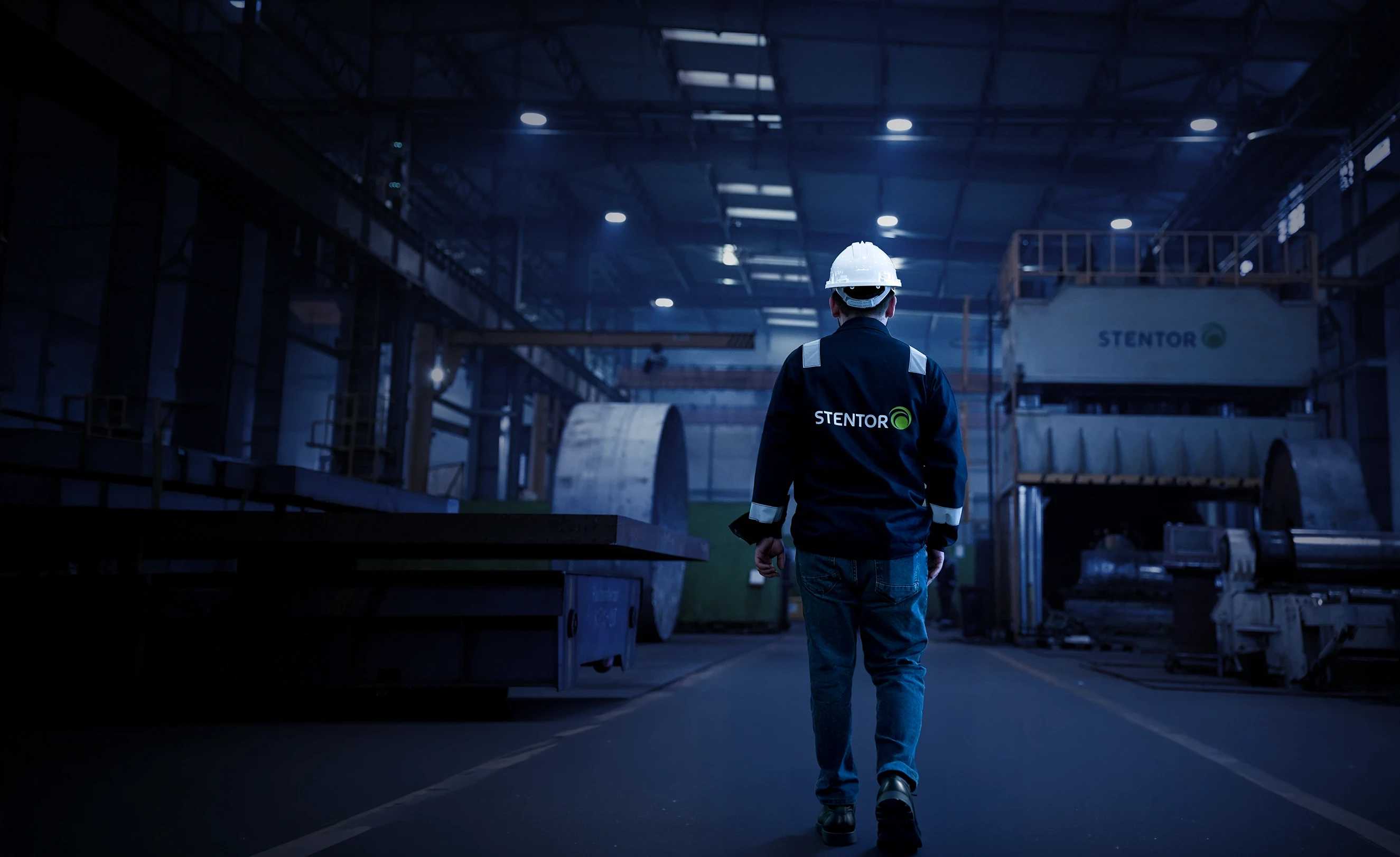 Muncitor purtând o cască de protecție și o jachetă cu inscripția STENTOR într-o fabrică industrială cu utilaje mari.