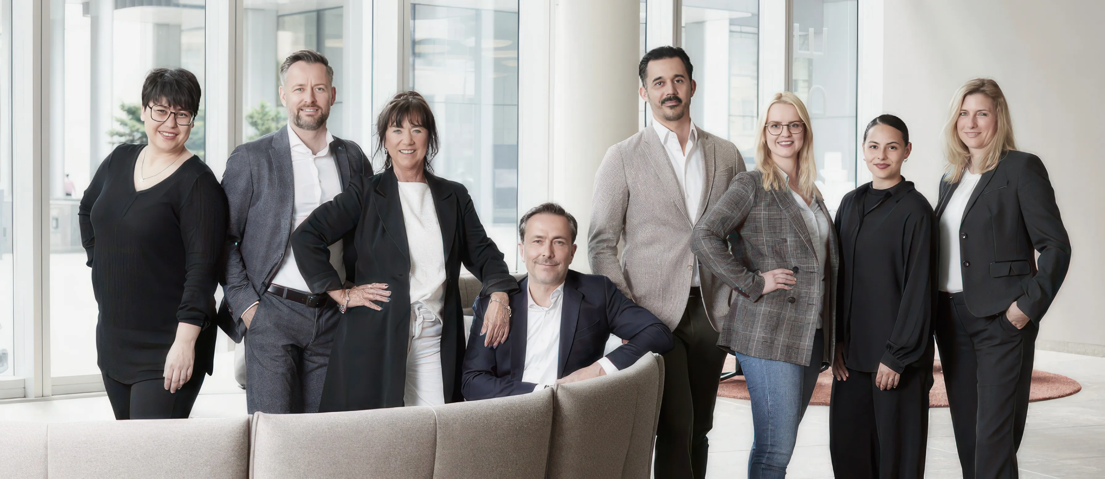 Acht Personen in Büro-Outfits posieren lächelnd vor großen Fenstern in modernem Gebäude.