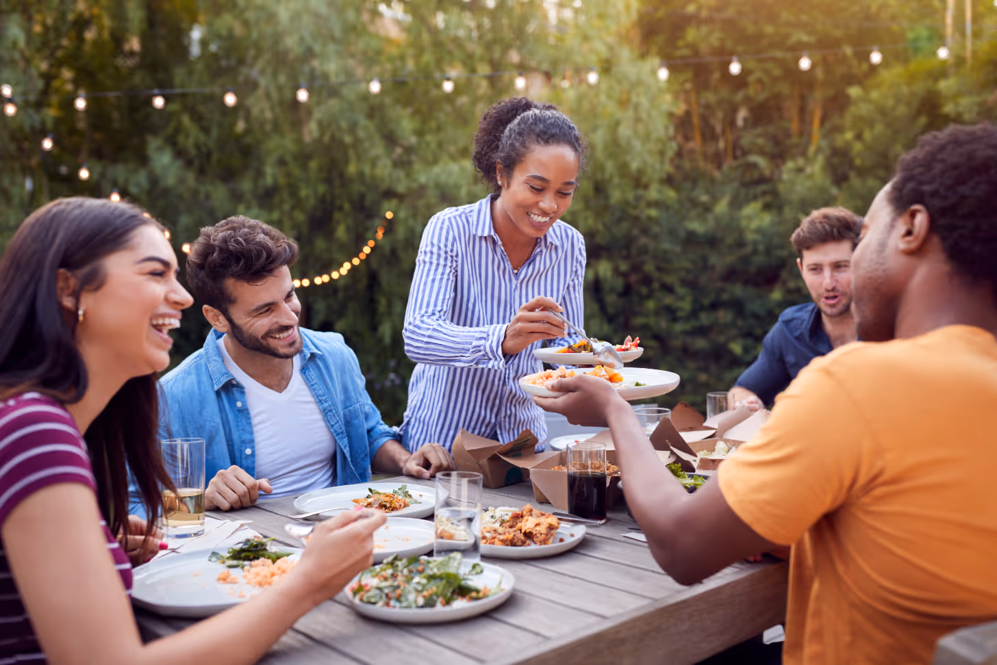 Multi-Cultural Friends At Home Sitting At Table Enjoying Food At Summer Garden Party stock image