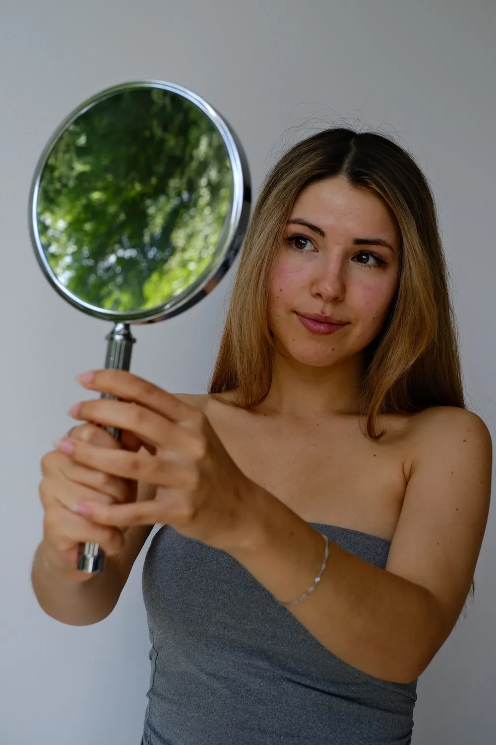 Frau mit langem Haar hält einen runden Handspiegel und betrachtet das Behandlungsergebniss ihrer Behandlung bei Skinthetics
